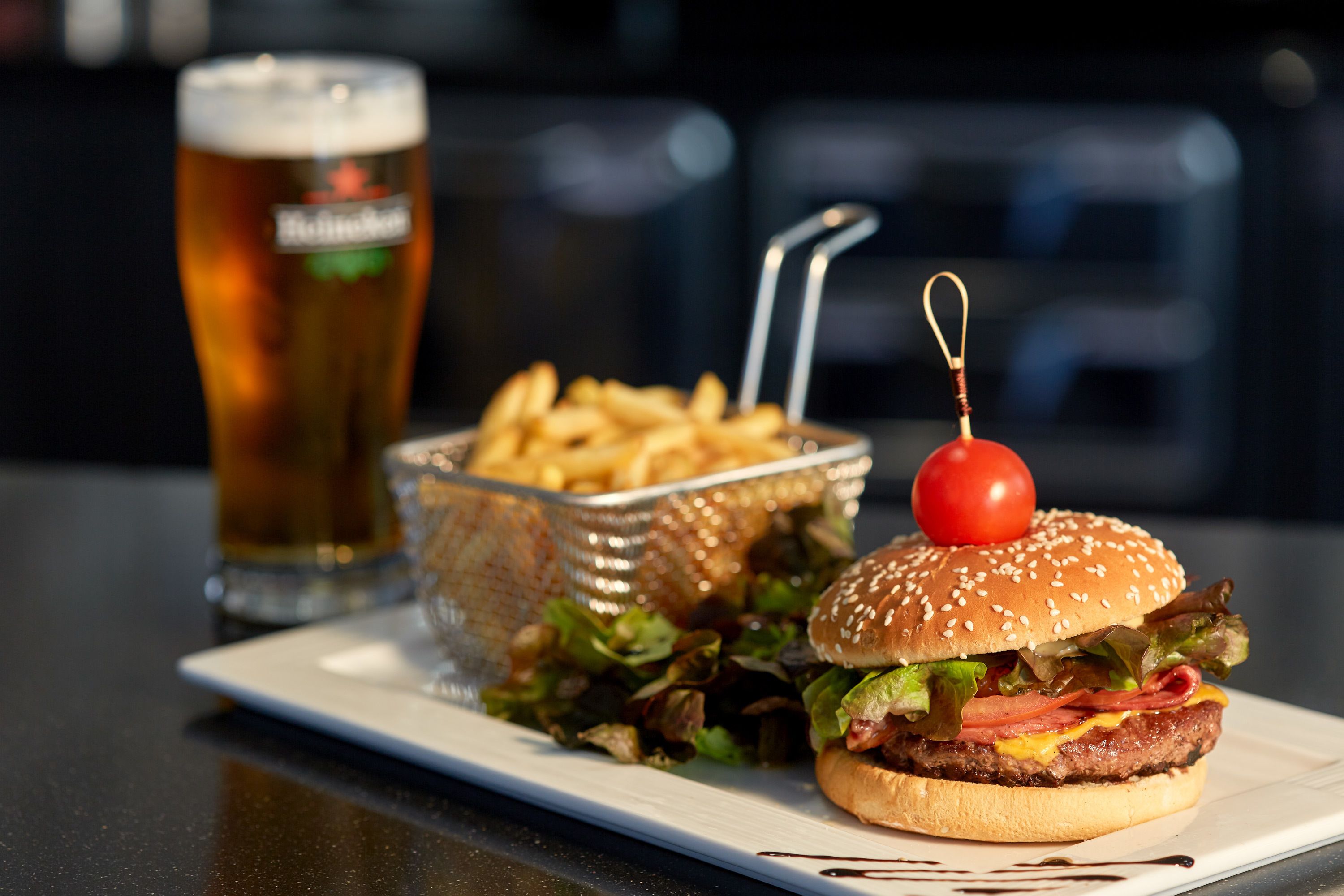 Un hamburger con una ciliegina sulla sommità, un bicchiere di birra e un cestino di patatine fritte su un piatto bianco.