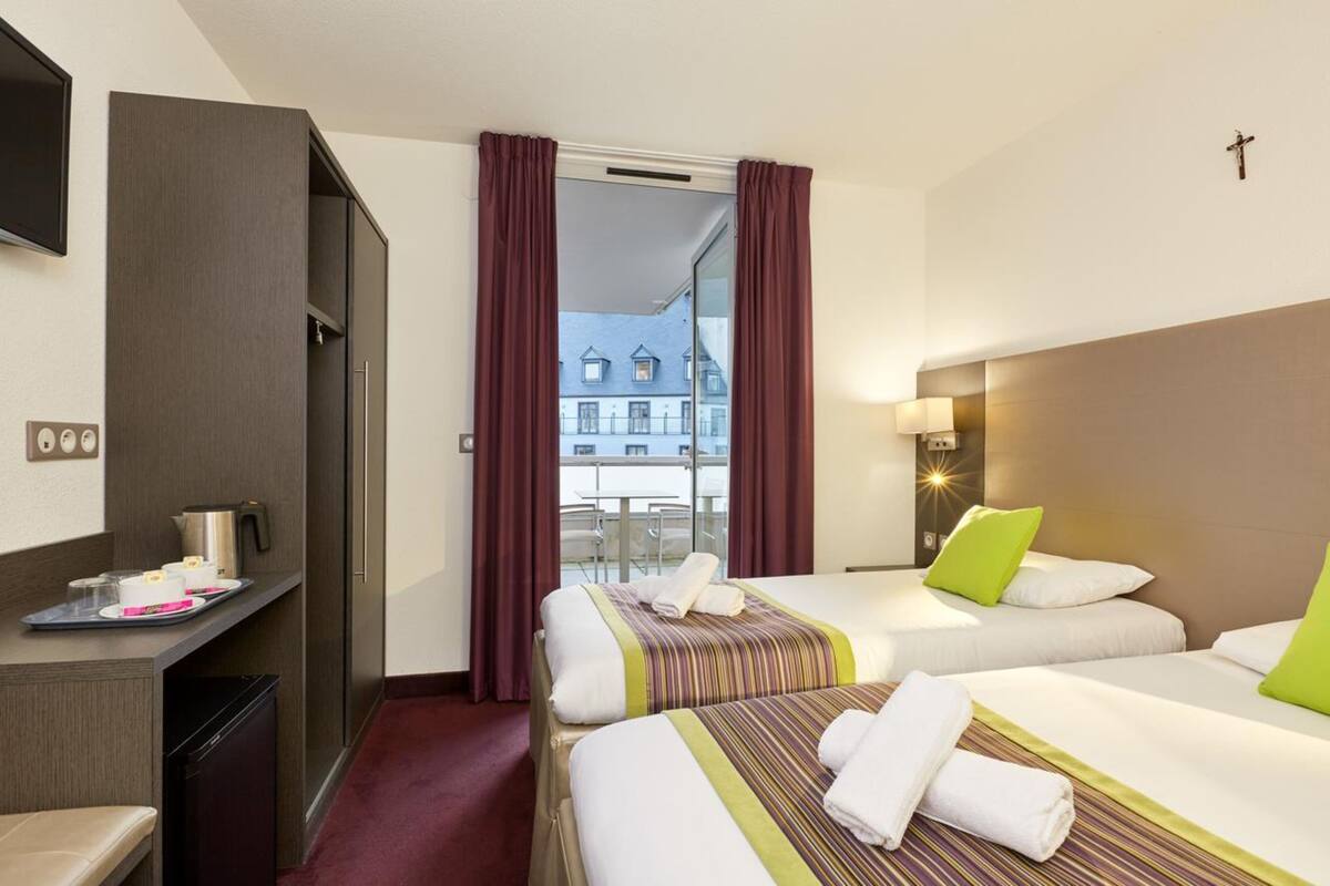 A hotel room with two beds, a desk with a coffee maker, a view of buildings outside, and a cross on the wall.