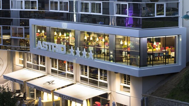 A modern hotel with a glass facade, outdoor seating area, and visible restaurant interior. - Hôtel Astrid (Lourdes)