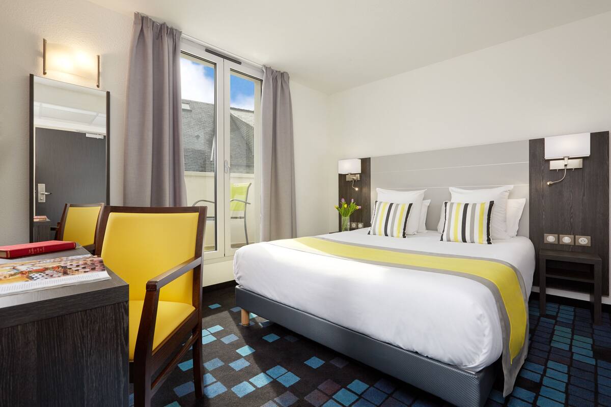 A hotel room with a bed, a desk with a chair, a mirror, and a window with curtains.