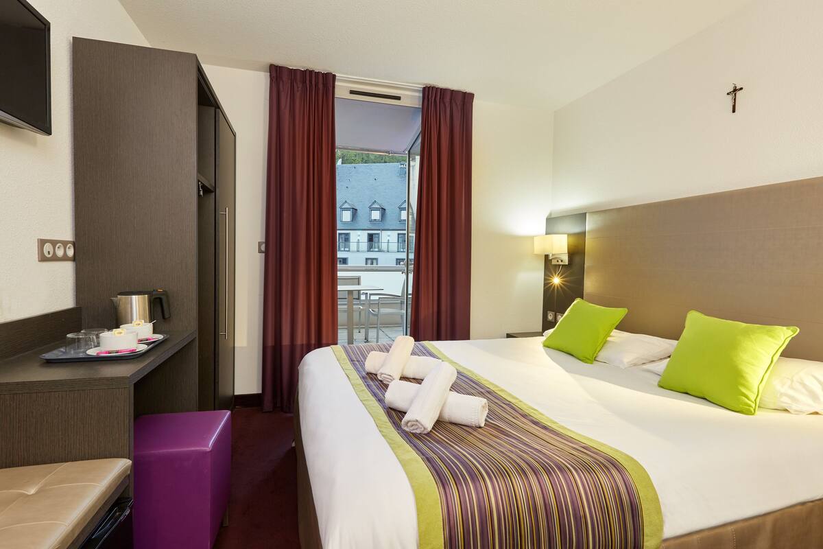 A hotel room with a bed, a desk with a kettle, a chair, and a view of buildings through the window.