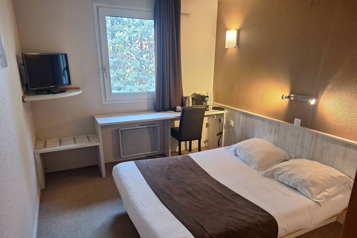 A hotel room with a bed, a desk, a chair, a television, a window with curtains, and wall-mounted lights.