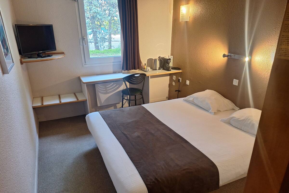 A hotel room with a bed, a desk with a chair, a television, and a window with curtains.