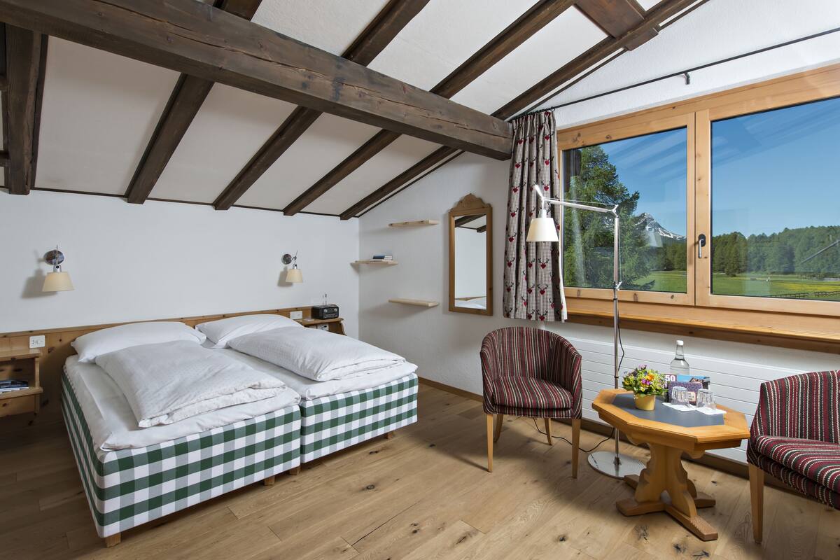 Ein Zimmer mit einem Bett, einem Sessel, einem Tisch, einer Lampe, einem Fenster mit Aussicht und einem Spiegel.