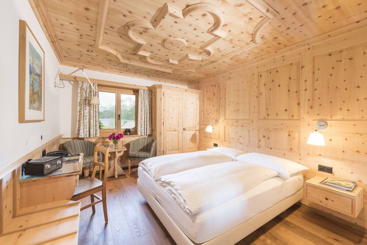 Ein Zimmer mit Holzboden, einem Bett, einem Schreibtisch und einem Sessel. Es gibt ein Fenster mit Vorhängen, eine Lampe und ein Gemälde an der Wand.