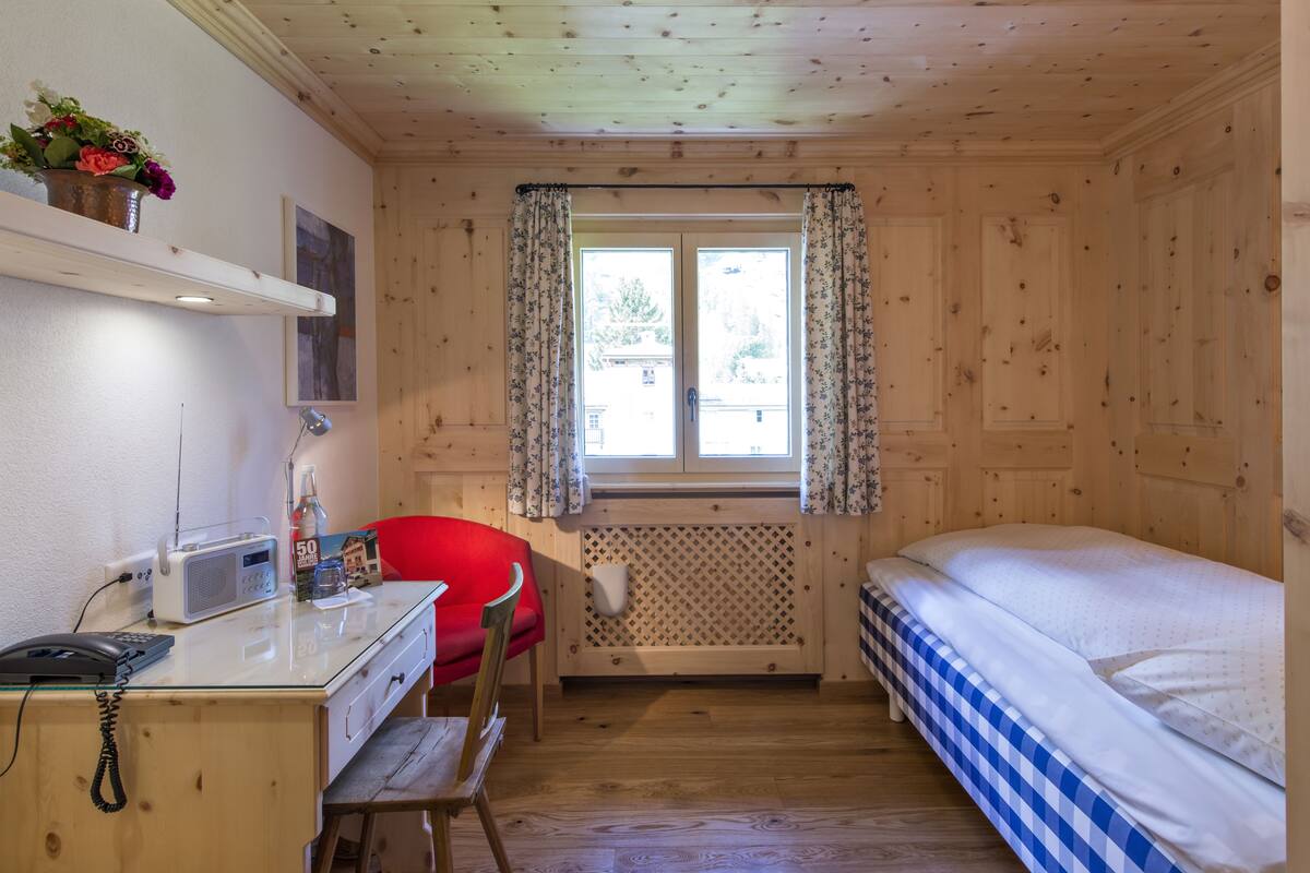 Ein Zimmer mit Holzvertäfelung, einem Bett mit blau-weißer Bettwäsche, einem Schreibtisch mit Telefon und Radio, einem roten Sessel und einem Fenster mit blumigem Vorhang.