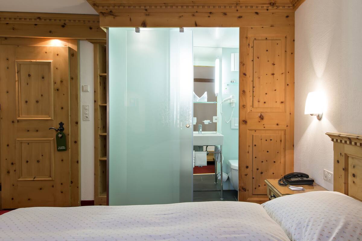Ein Hotelzimmer mit Holztür, Duschabtrennung aus Glas, einem Bett mit weißer Bettdecke, einem Nachttisch mit Lampe und einem kleinen Schreibtisch mit Telefon.