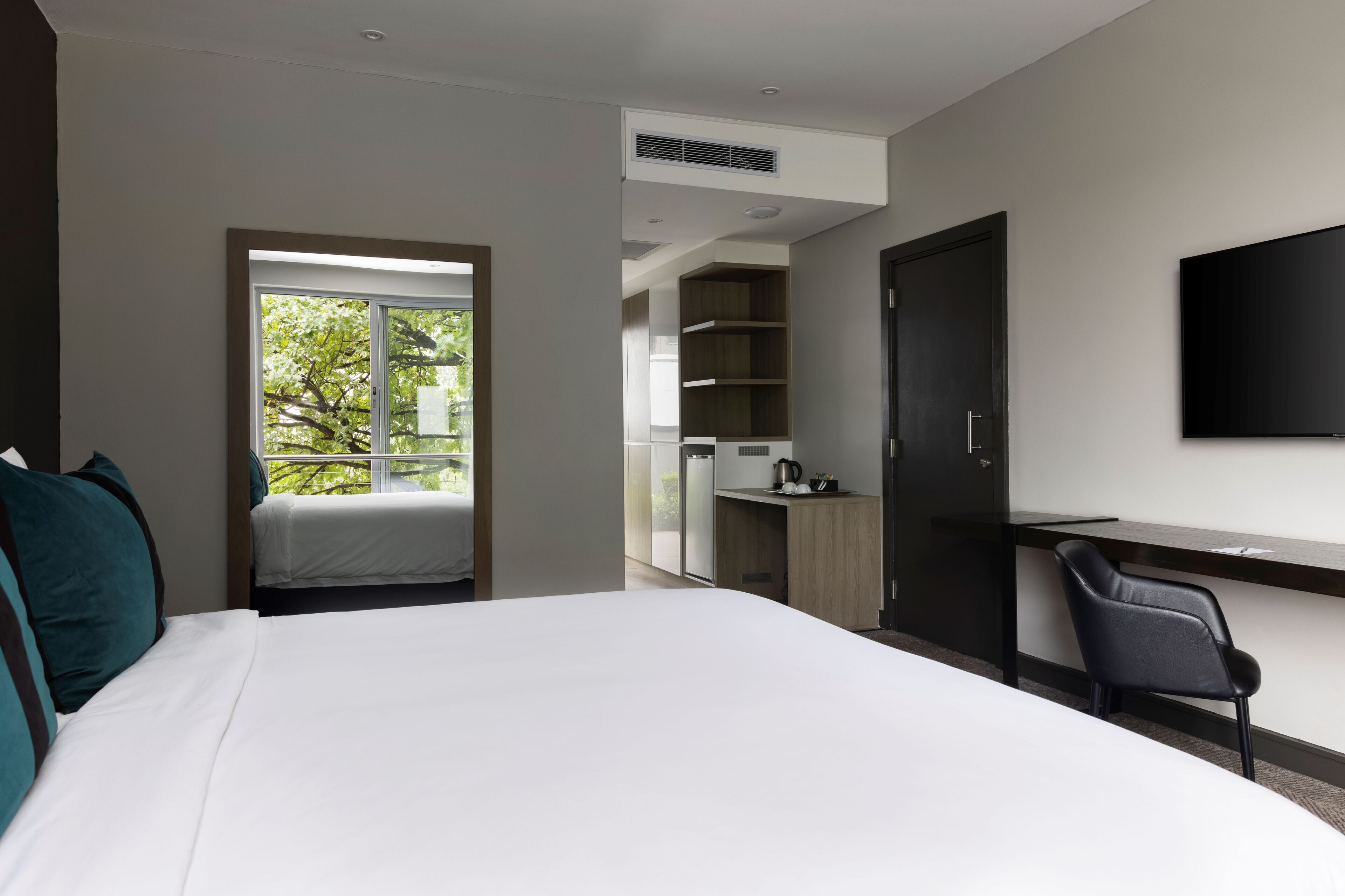 Habitación de hotel moderna con una cama grande, un escritorio, una silla, un televisor y un balcón con vistas a árboles.