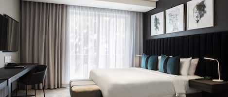 A modern hotel room with a bed, desk, and chair, a TV, and artwork on the wall.