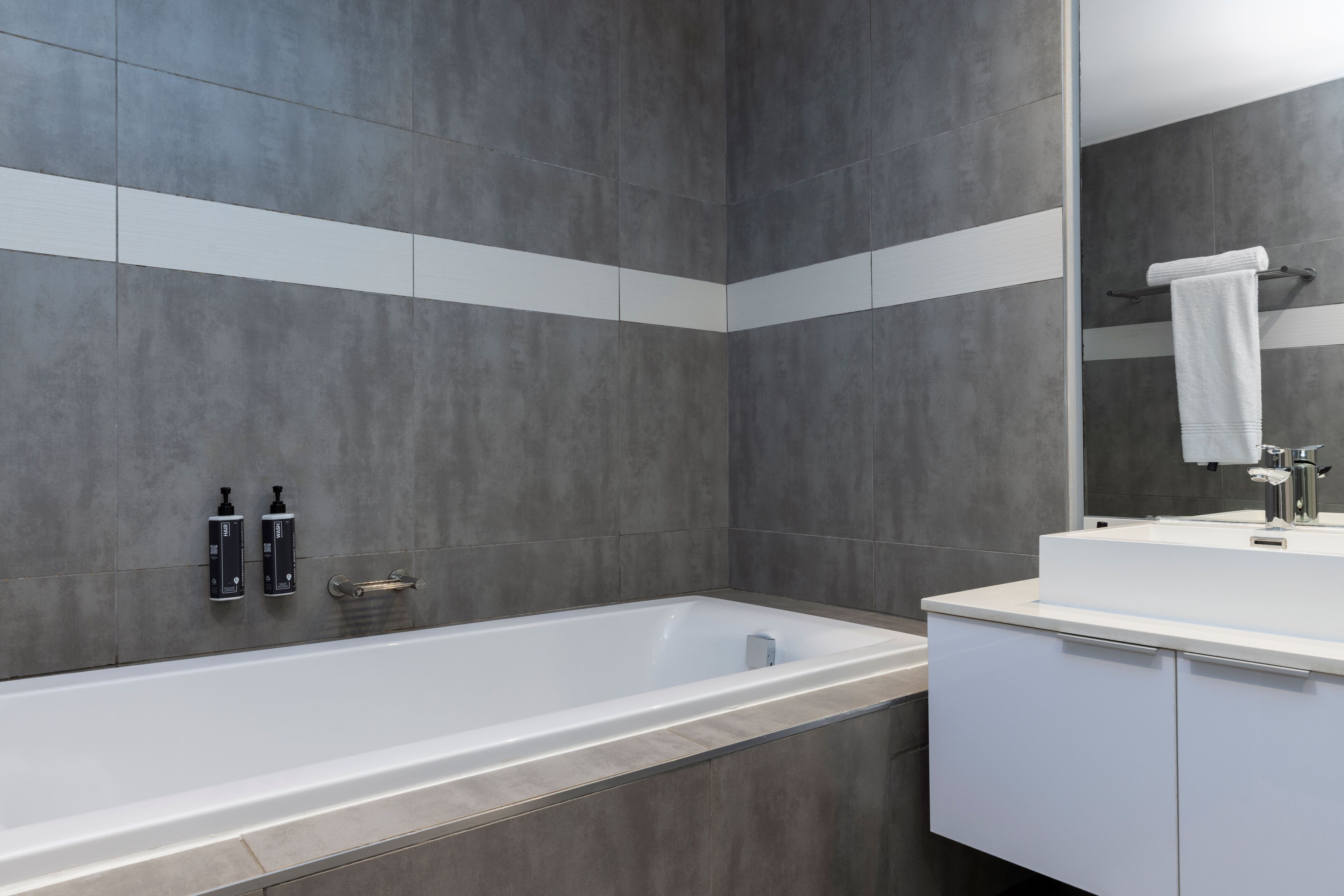 Un bagno moderno con una vasca da bagno ampia, un mobile lavabo bianco con specchio e pareti rivestite di piastrelle grigie.