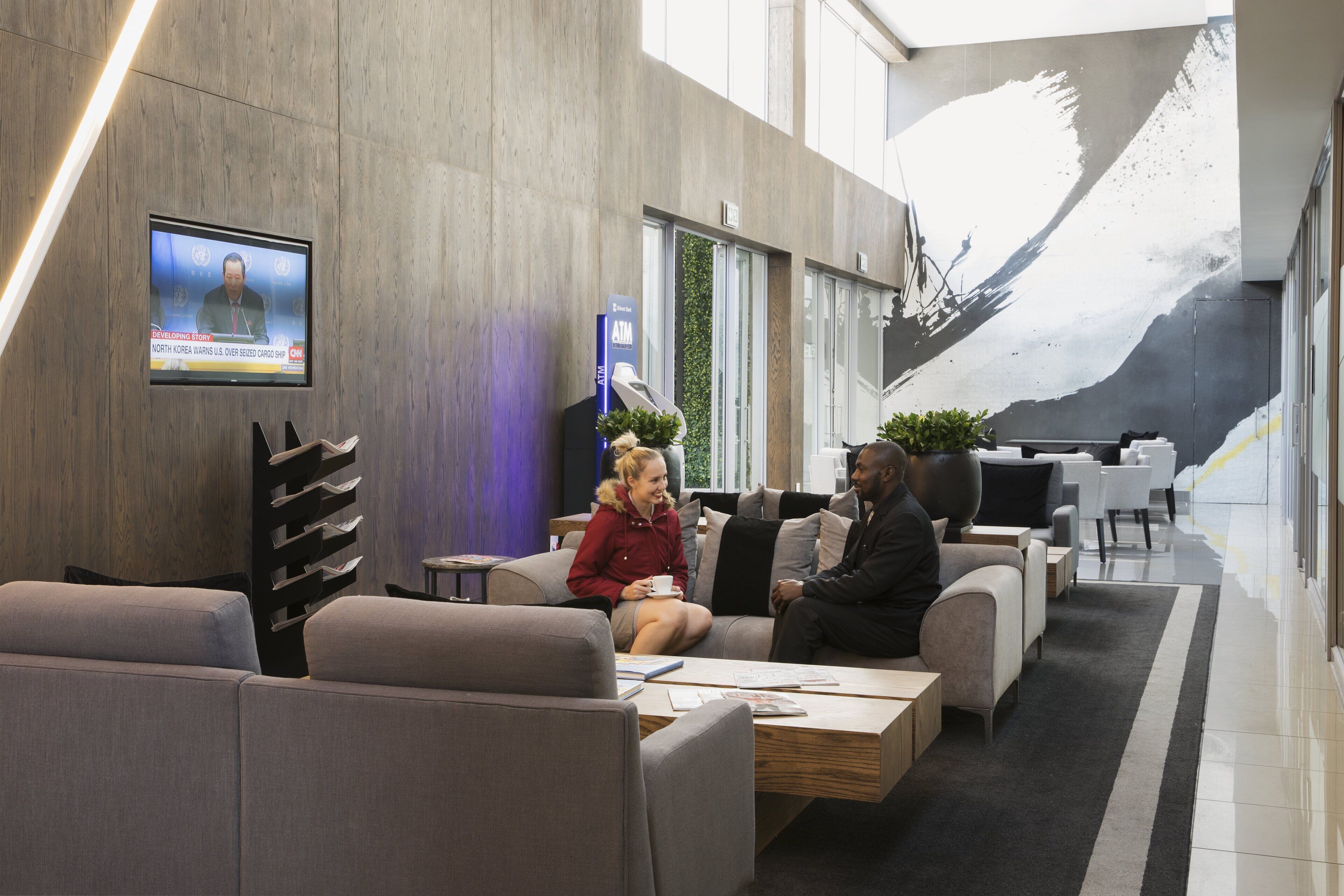A modern lobby with a large screen displaying a news broadcast, comfortable seating areas, and a contemporary art piece on the wall.