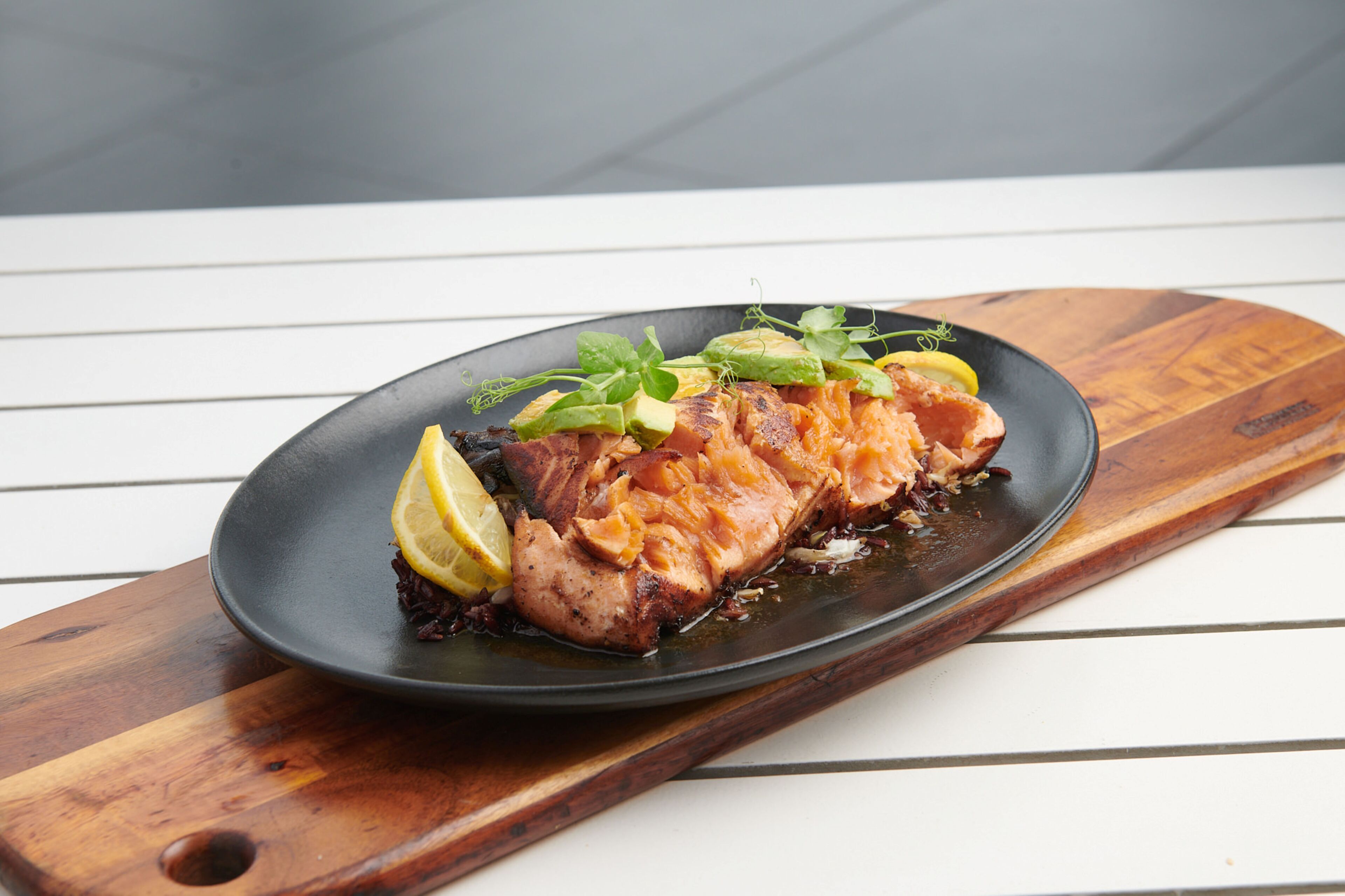 Un plato de salmón con rodajas de limón y verduras sobre un plato negro sobre una tabla de madera.