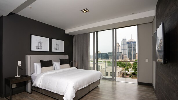 Une chambre moderne dotée d’un grand lit, d’une table de chevet, d’un téléviseur à écran plat et offrant une vue sur la ville grâce à de grandes fenêtres du sol au plafond.