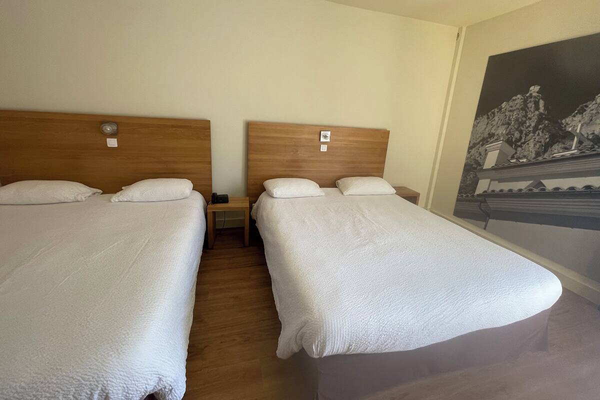 Une chambre d’hôtel avec deux lits simples, des têtes de lit en bois, du linge de lit blanc, une petite table de chevet avec un téléphone et une lampe fixée au mur.