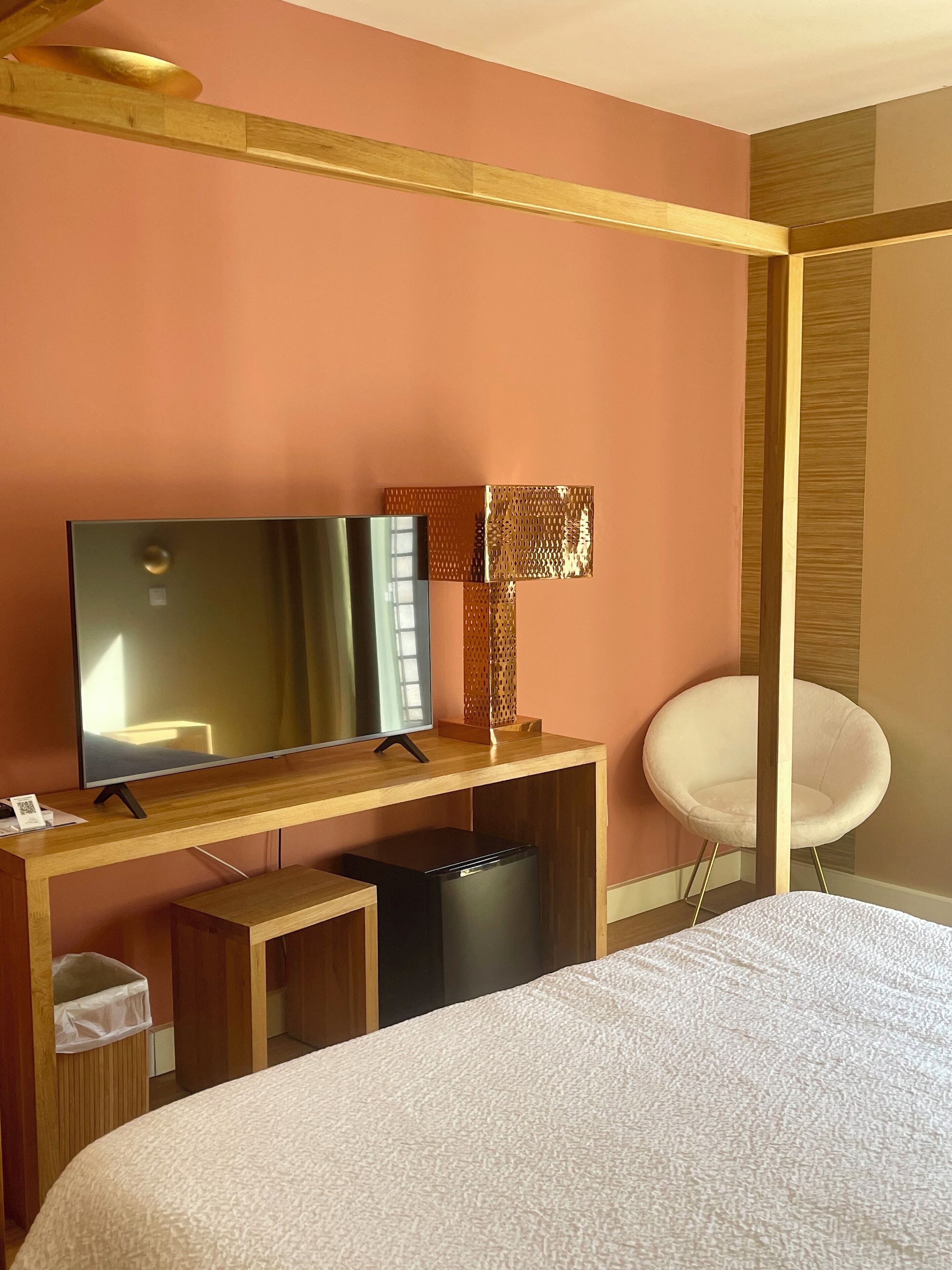 A hotel room with a bed, a TV on a wooden stand, a small black trash bin, and a white chair.