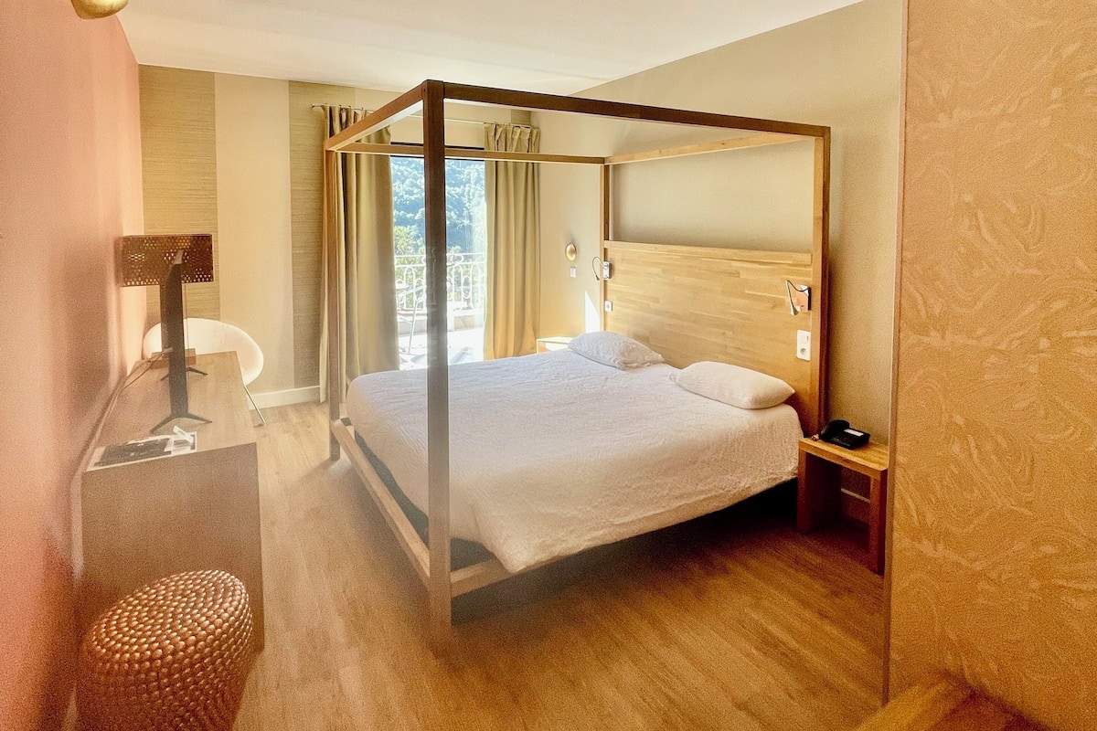 Un lit à baldaquin dans une chambre d’hôtel, avec un plancher en bois, un bureau avec une lampe et une vue sur l’extérieur par la fenêtre.