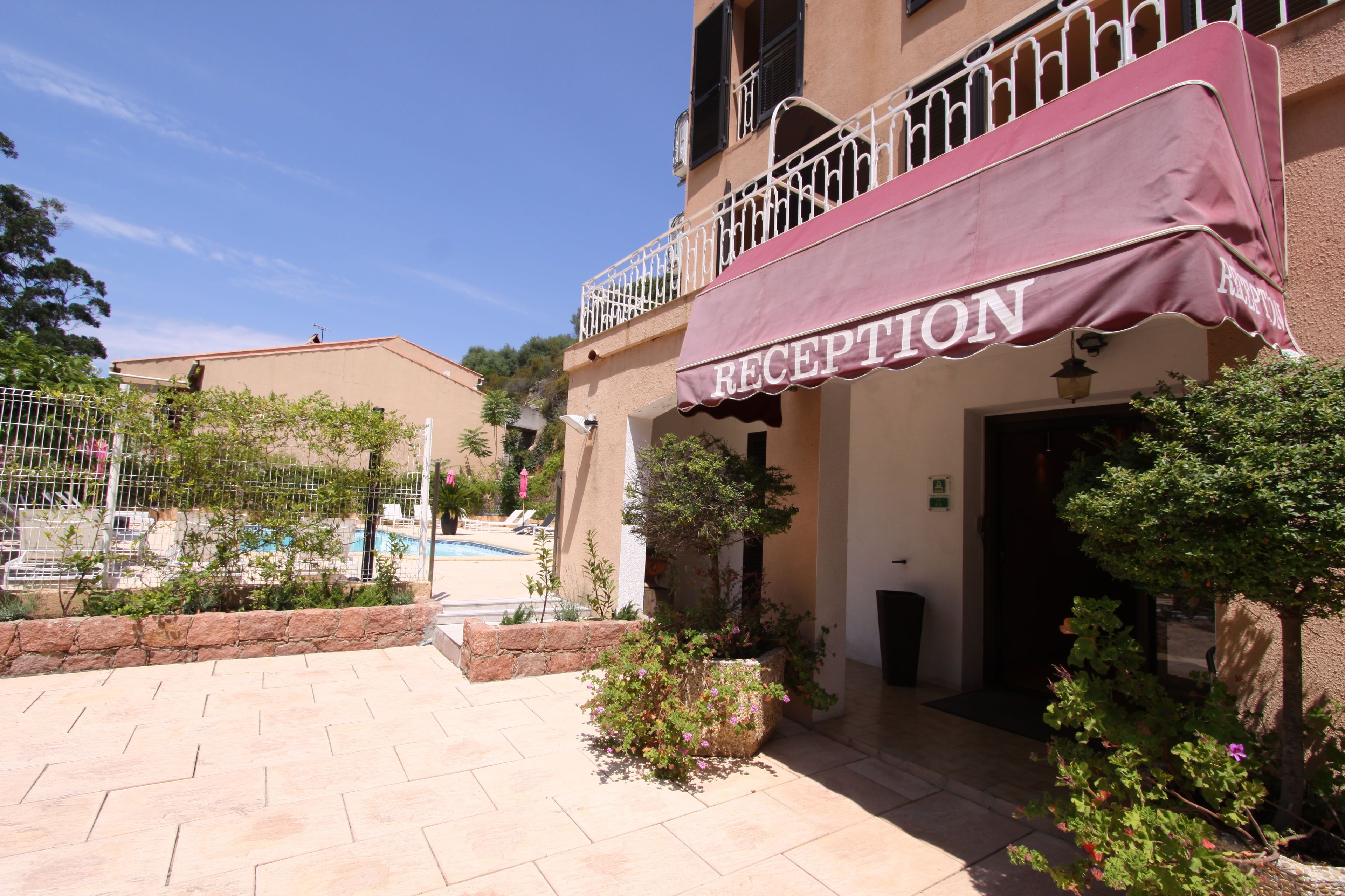 A hotel reception with a balcony, a pool in the background, and a stone pathway.