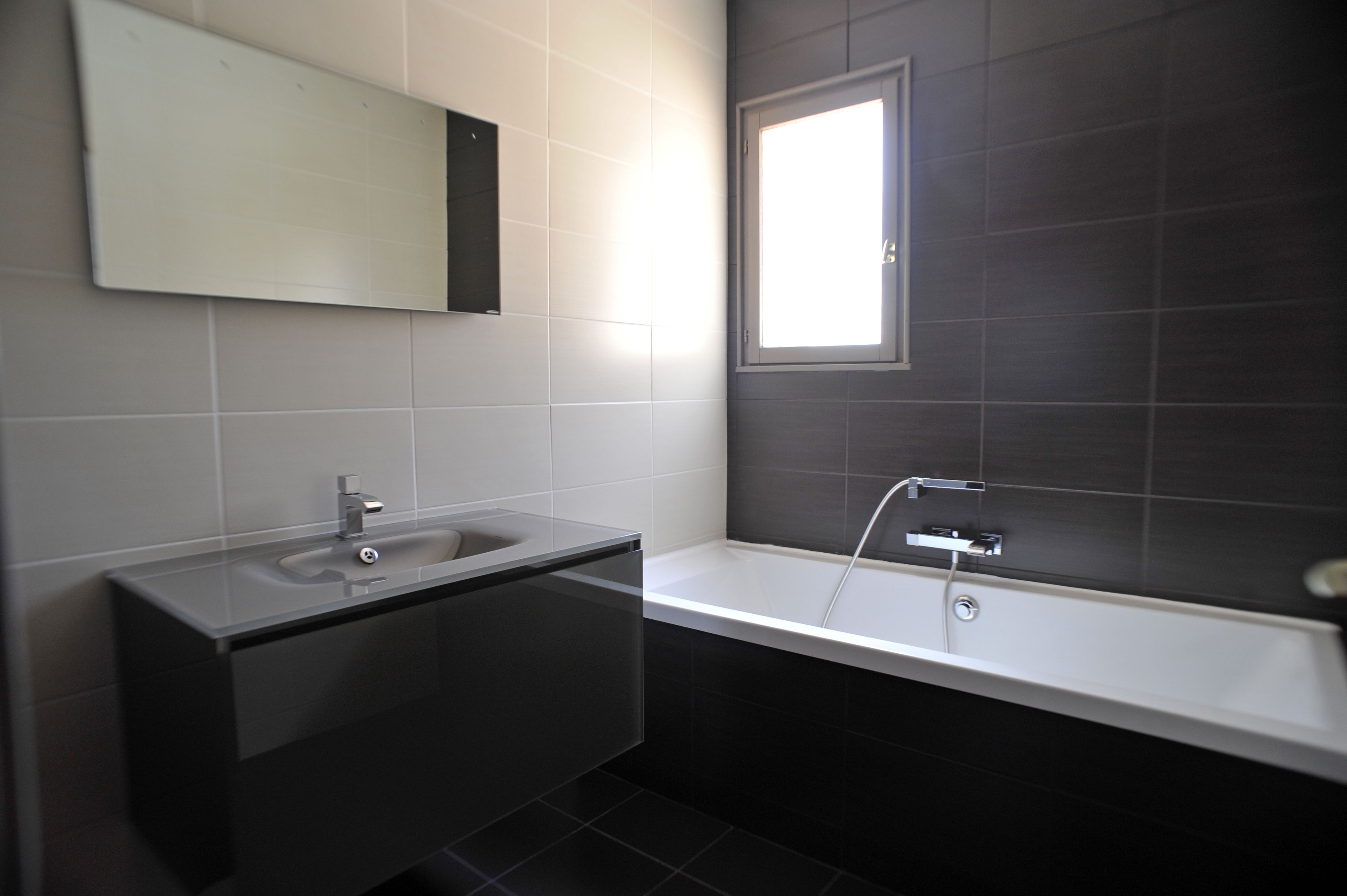 Un baño moderno con pared de azulejos oscuros, un espejo grande, un lavamanos blanco y una bañera blanca.