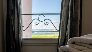 Een raam met een metalen balustrade met uitzicht op een strand, een opgevouwen handdoek op de vloer, en gordijnen aan weerszijden.