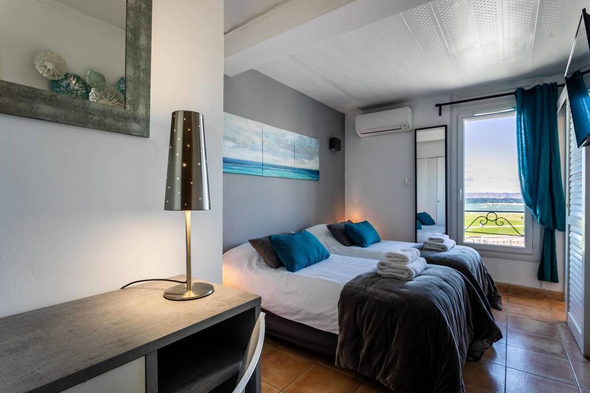 A hotel room with two beds, a desk, a lamp, a painting, an air conditioning unit, and a window with curtains.
