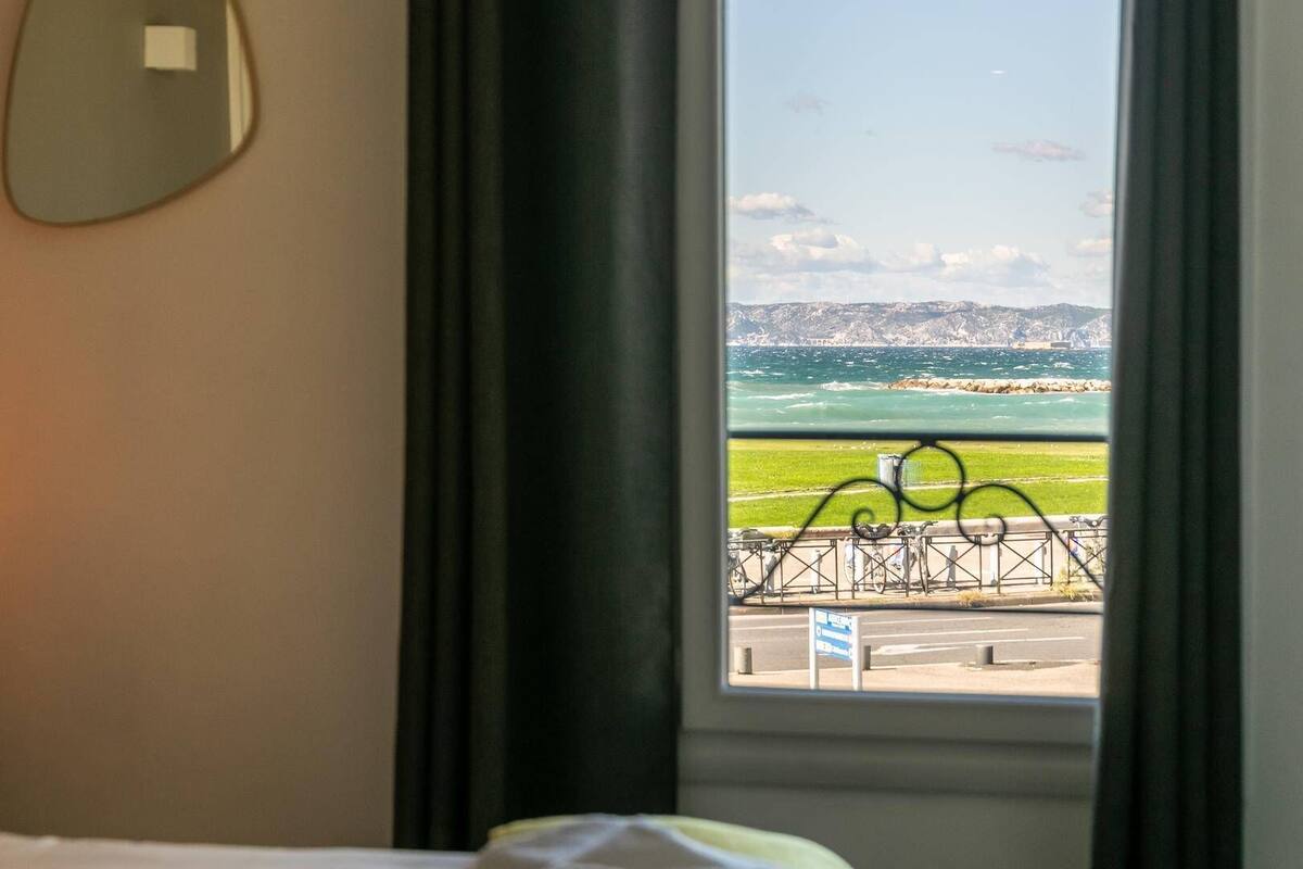 A hotel room with a window view of a coastal landscape, including a grassy area and a metal fence.