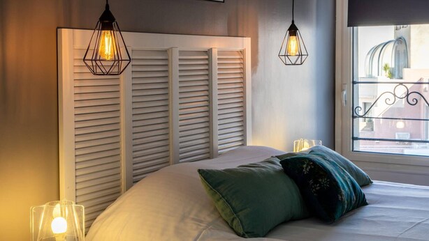Een slaapkamer met een bed, twee bedlampen en een raam met uitzicht op gebouwen.