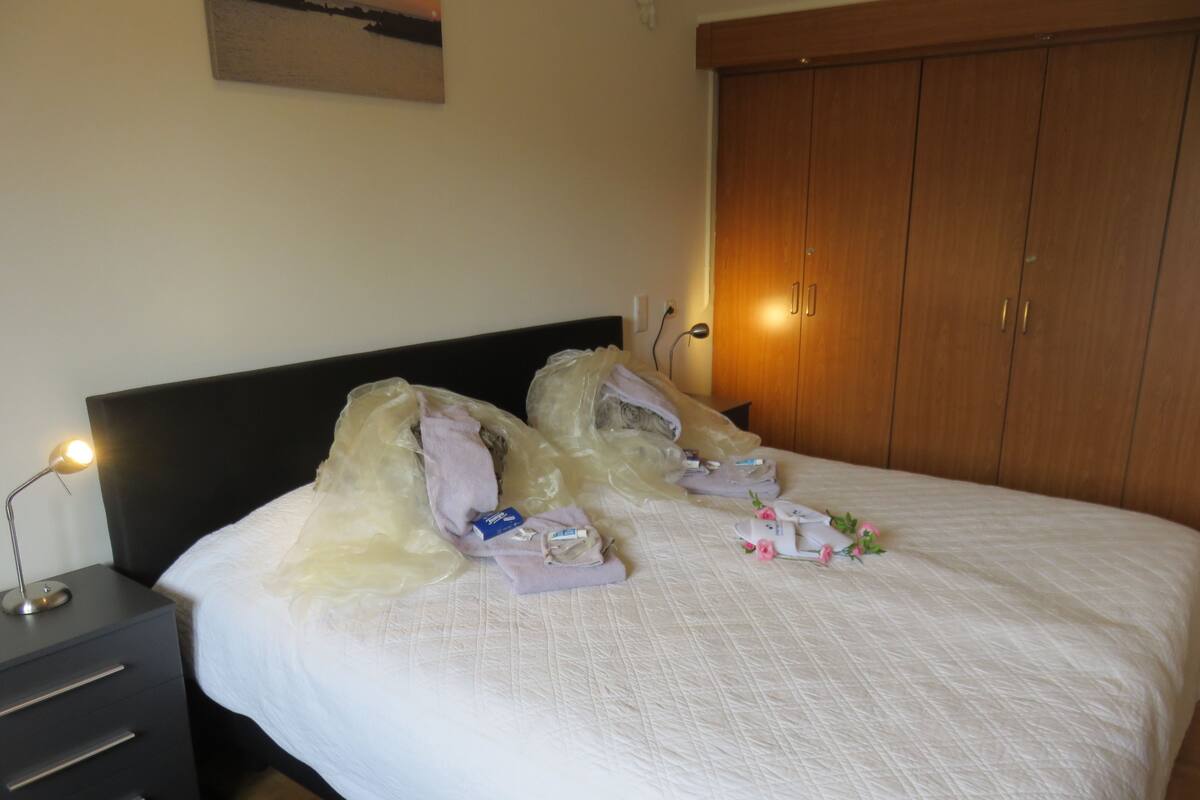 A neatly made bed with a white duvet, a wooden headboard, and a wooden wardrobe in the background.