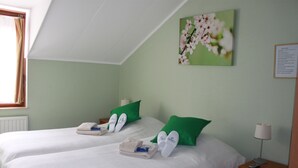 A hotel room with two beds, green pillows, white linens, a painting of flowers, a lamp, and a nightstand with a remote control.