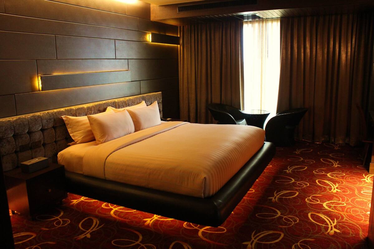 Habitación de hotel con una cama grande, un cabecero con iluminación integrada, una alfombra roja estampada y una ventana con cortinas.