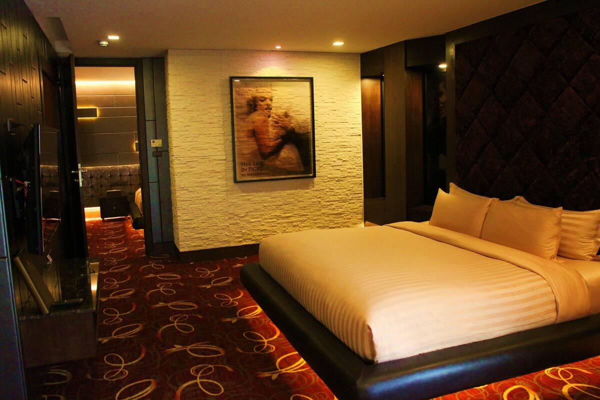 Habitación de hotel con una cama grande, una alfombra roja con estampado, un cuadro en la pared y un cabecero oscuro.