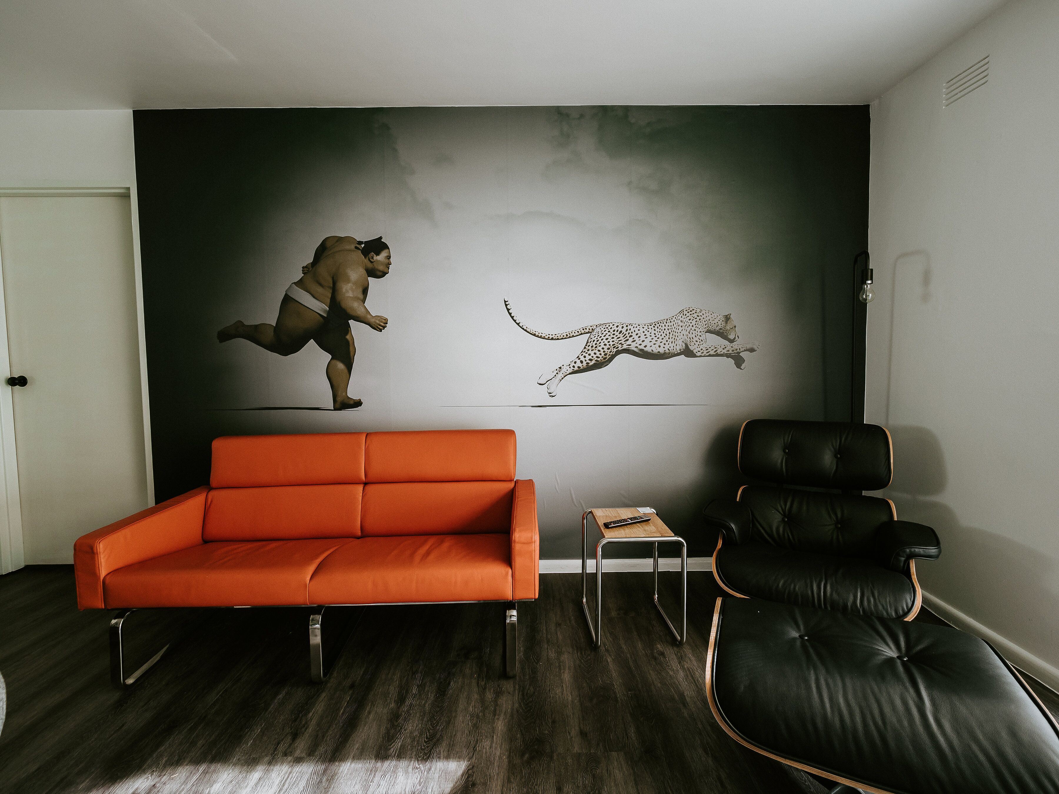 Uma sala de estar moderna com piso de madeira escura, um sofá laranja, uma poltrona de couro preto e uma pequena mesa de madeira.