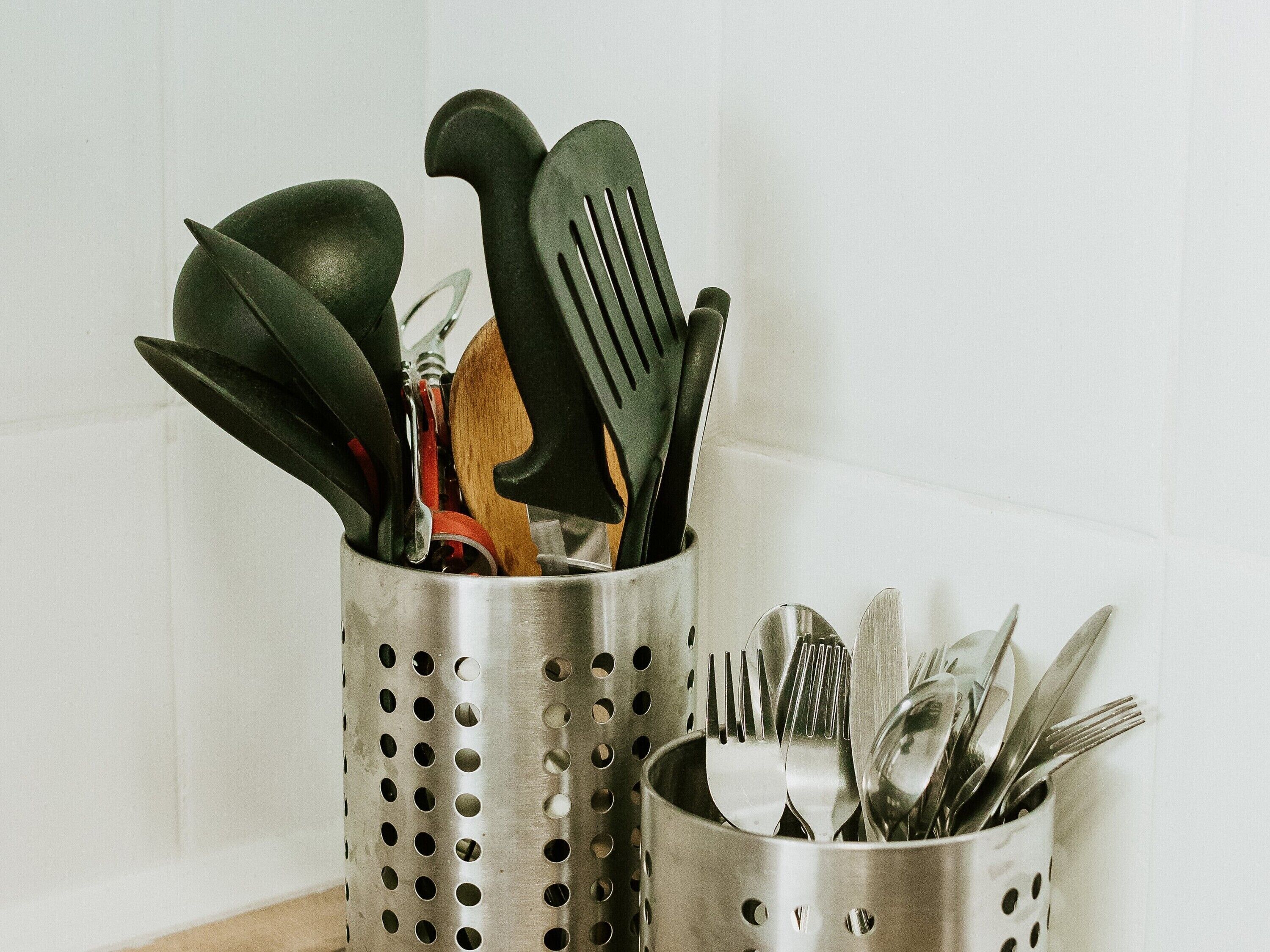 Two stainless steel utensil holders with holes, one containing a wooden spoon and the other holding forks and spoons.