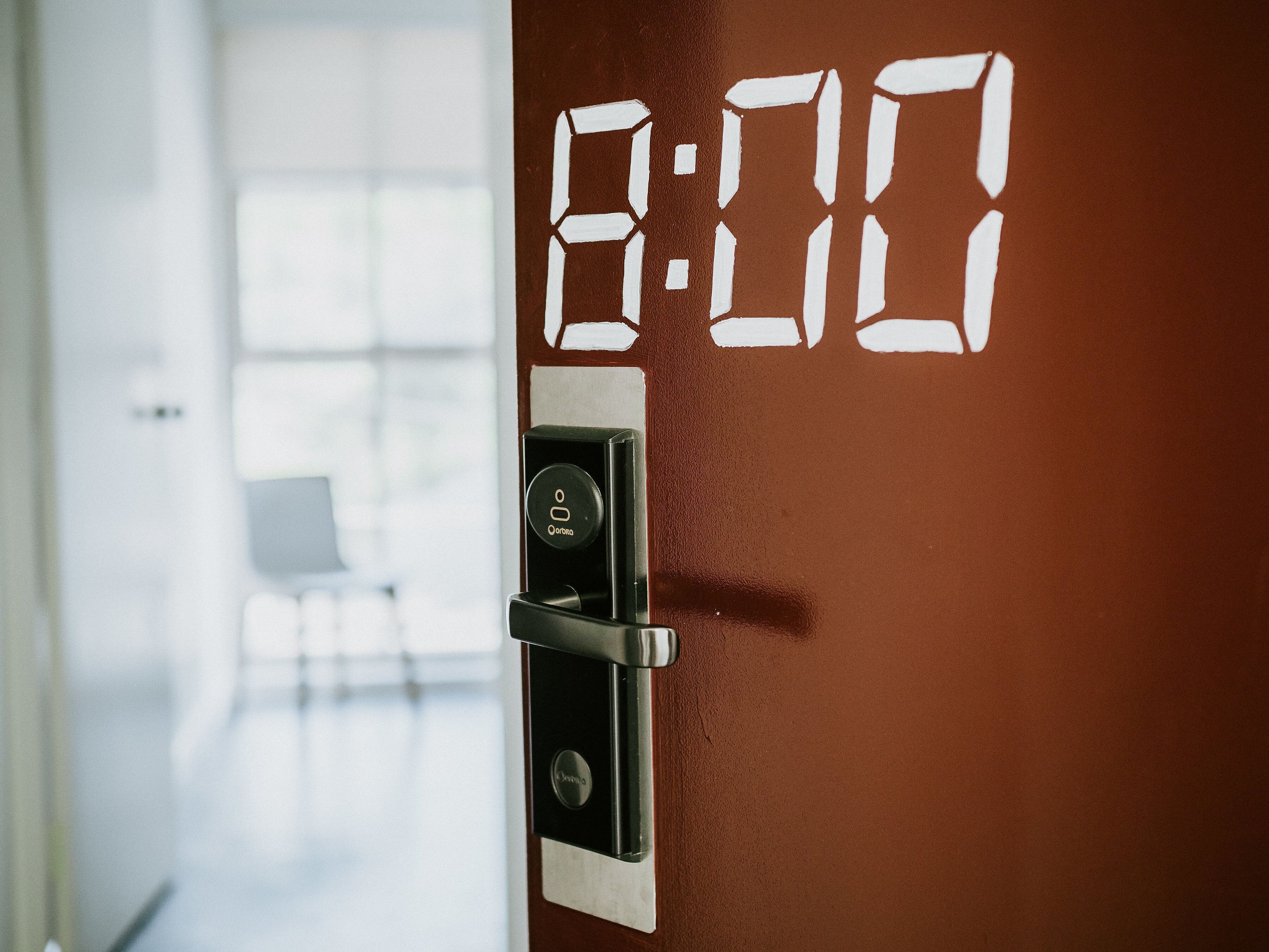 Un reloj digital en una puerta que muestra las 8:00, un lector de tarjetas de acceso y un interior moderno con una puerta de cristal y una silla al fondo.