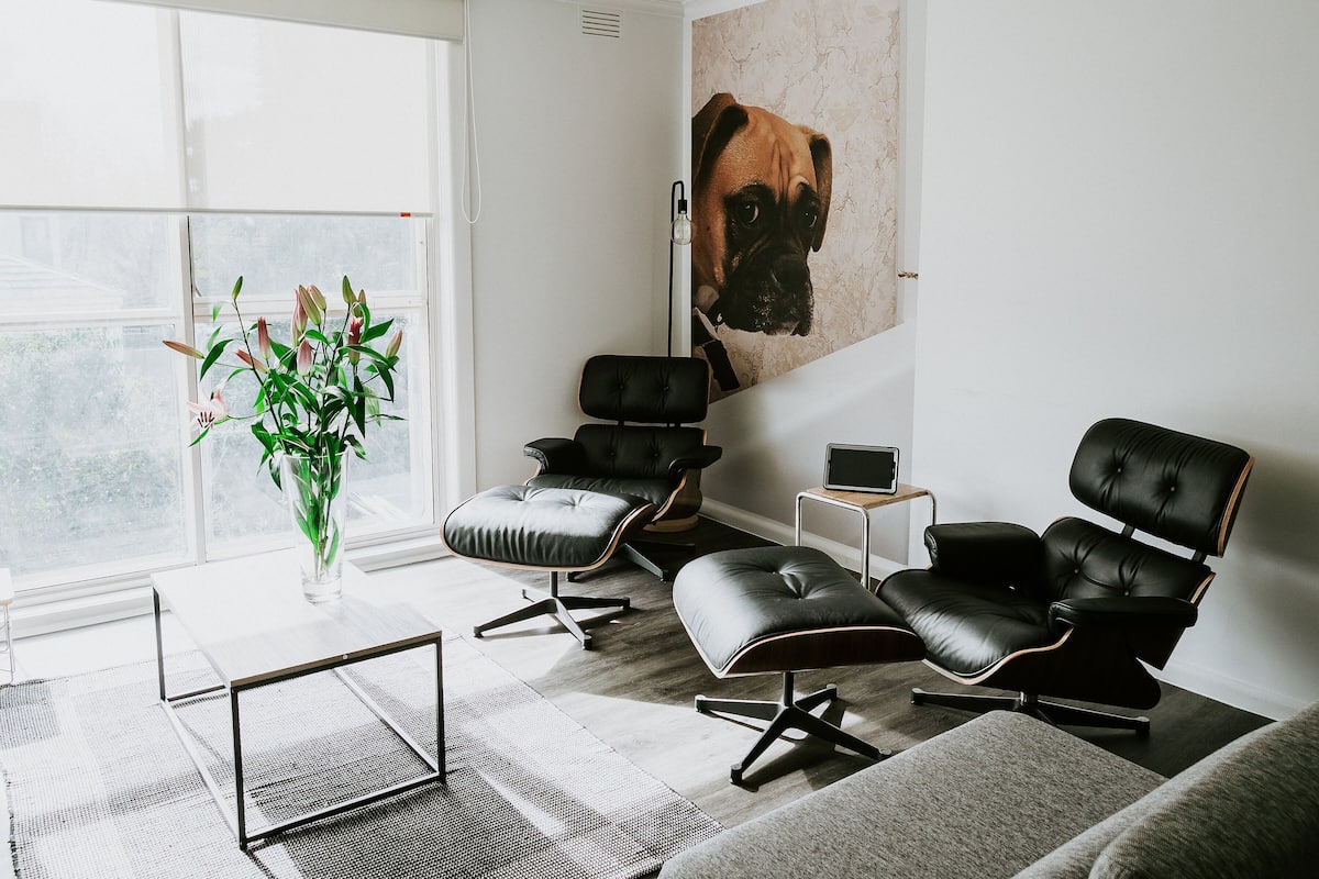 A modern living room with a large window, a black leather sofa, a black leather armchair, a small white table, and a potted plant.
