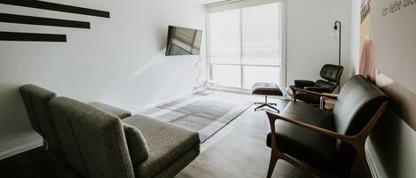 Una sala de estar moderna con un ventanal grande, un televisor de pantalla plana montado en la pared y una zona de estar con sofá y sillones.