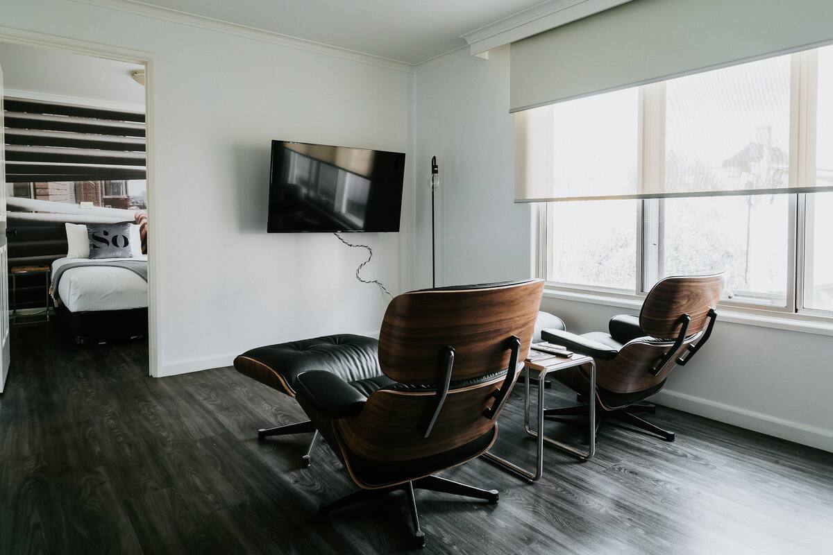 A modern room with a flat-screen TV mounted on the wall, two leather chairs, a small wooden table, and a view of the city through large windows.