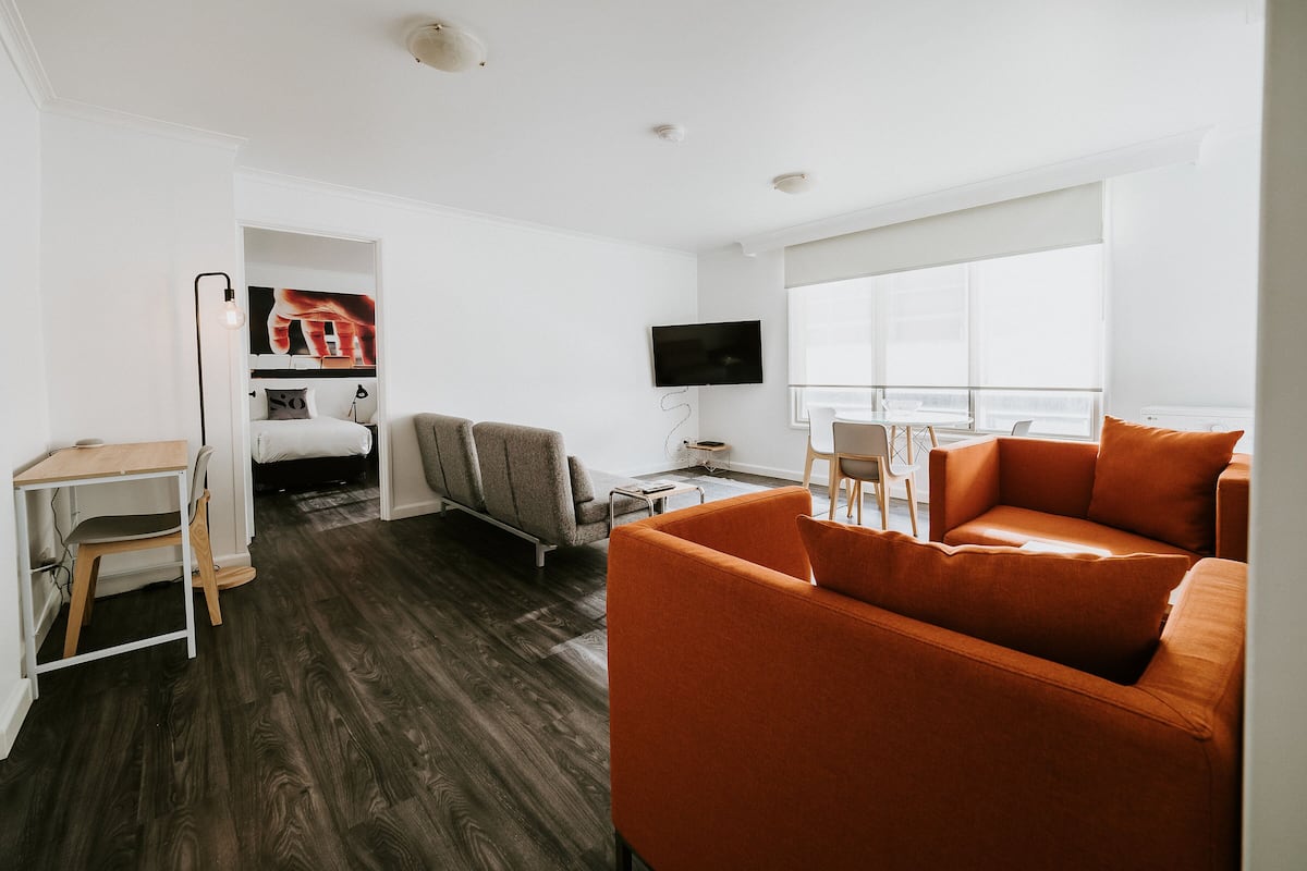 A modern hotel room with a wooden floor, a sofa, a desk with a chair, a bed, and a TV mounted on the wall.