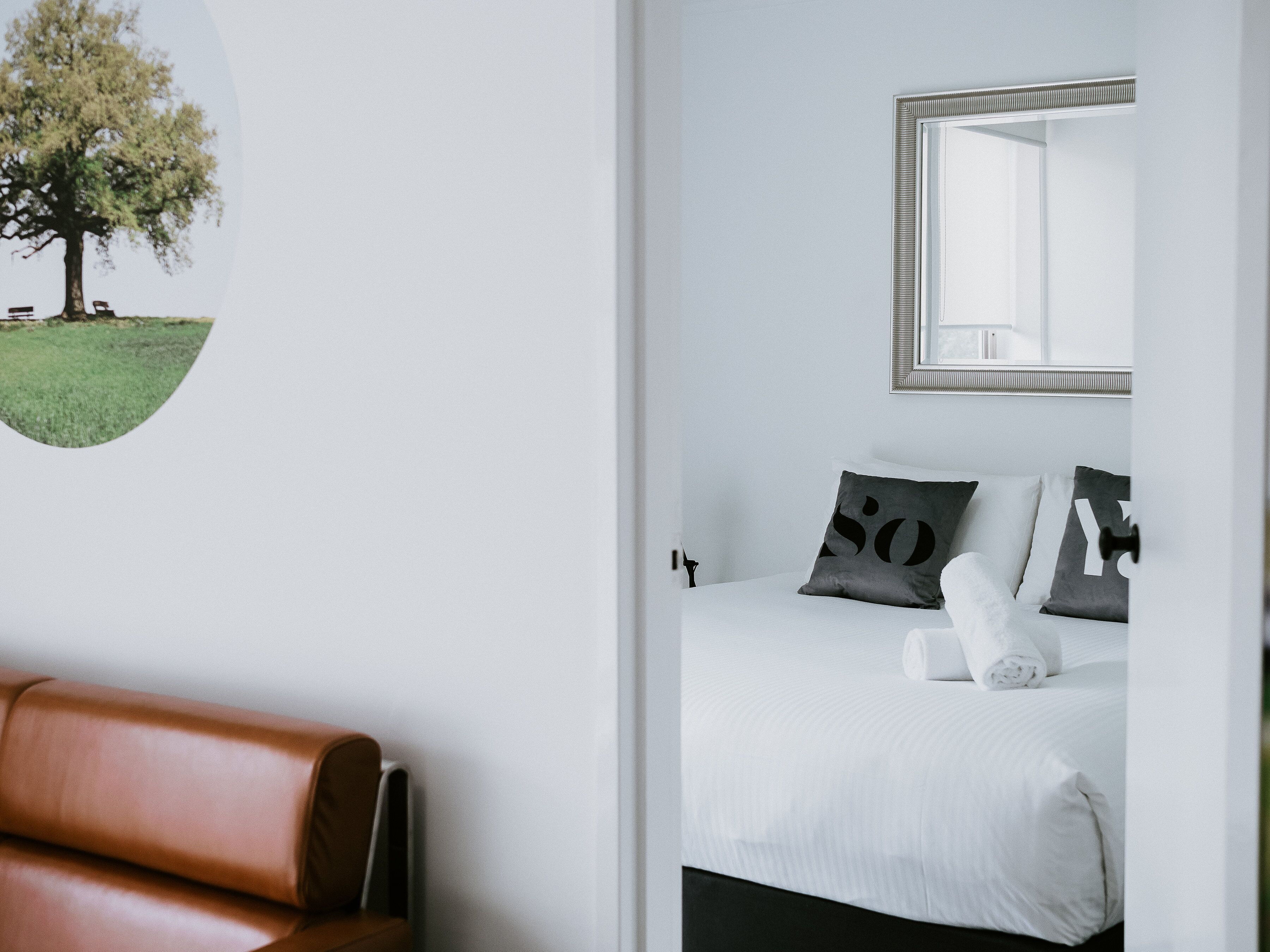 A hotel room with a bed, a brown leather chair, a wall-mounted mirror, and a wall art of a tree.