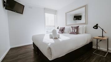 A modern hotel room with a large bed, a flat-screen TV mounted on the wall, a bedside table with a lamp, and a window providing a view of the city.