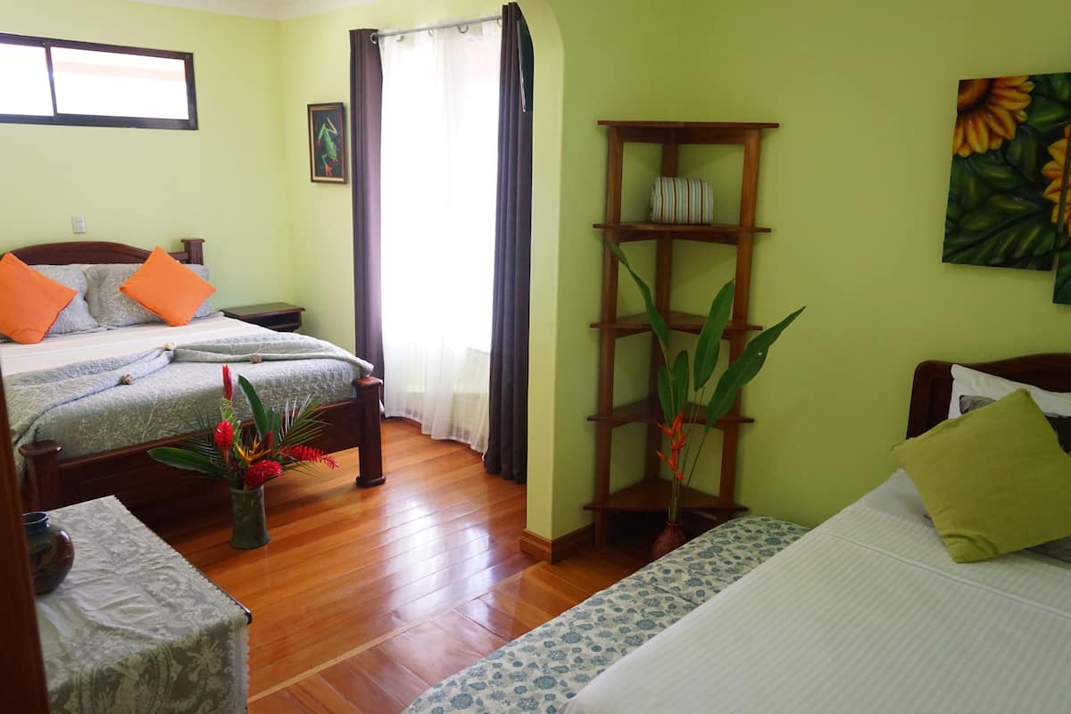A room with two beds, a wooden floor, a bedside table with a vase of flowers, a shelf with decorative items, and a painting on the wall.