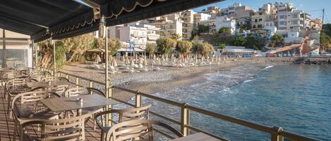 Un espace de restauration en plein air, avec des tables et des chaises, offrant une vue sur une plage et des bâtiments en arrière-plan.