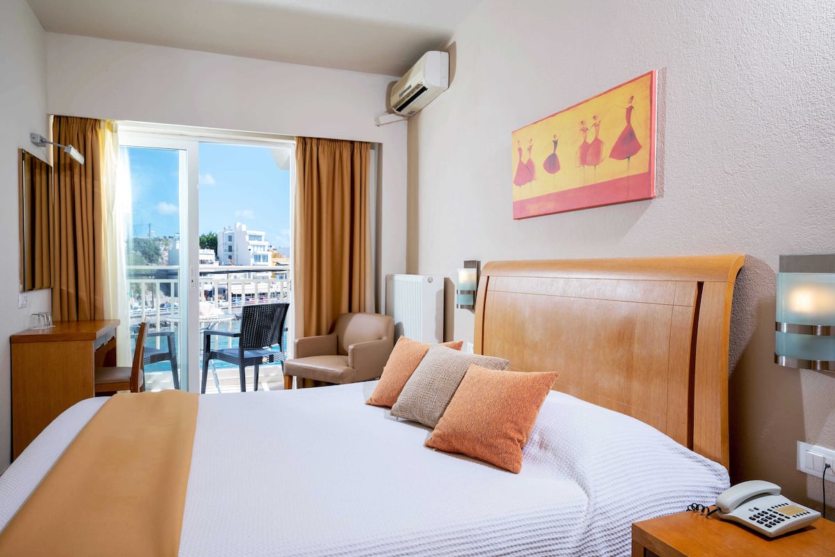 Ein Hotelzimmer mit einem großen Bett, einem Schreibtisch, einem Sessel und einem Balkon mit Blick auf Gebäude.