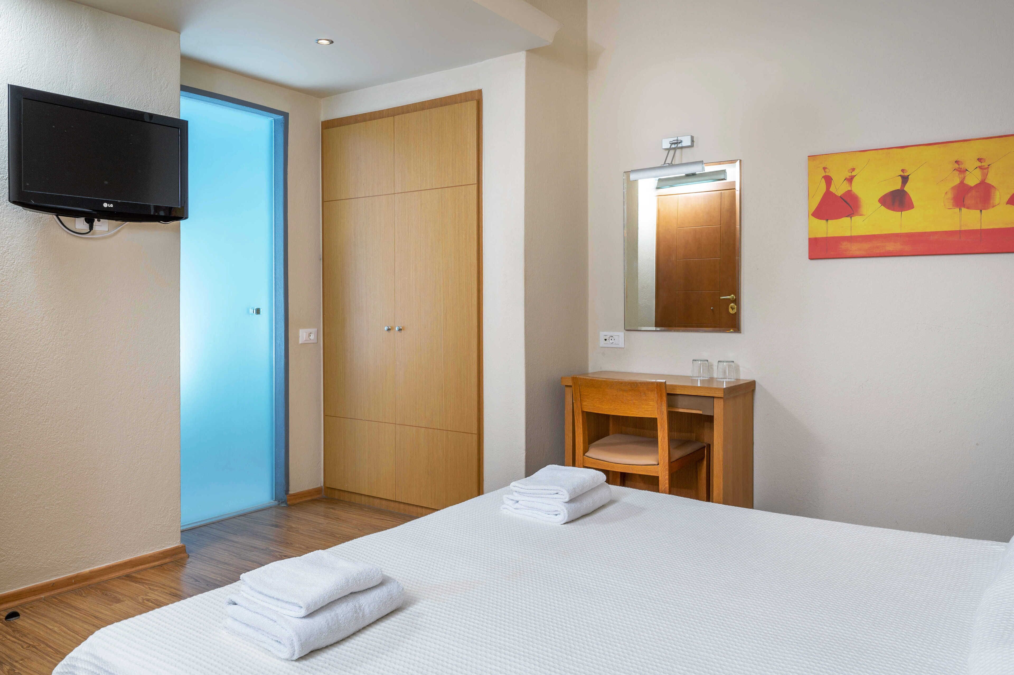 Habitación de hotel con cama, armario de madera, una mesita pequeña con espejo y televisor montado en la pared.