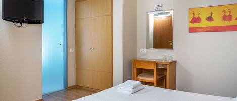 Habitación de hotel con cama, armario de madera, una mesita pequeña con espejo y televisor montado en la pared.