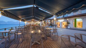 A waterfront restaurant with outdoor seating, a covered area, and a view of the sea and mountains.