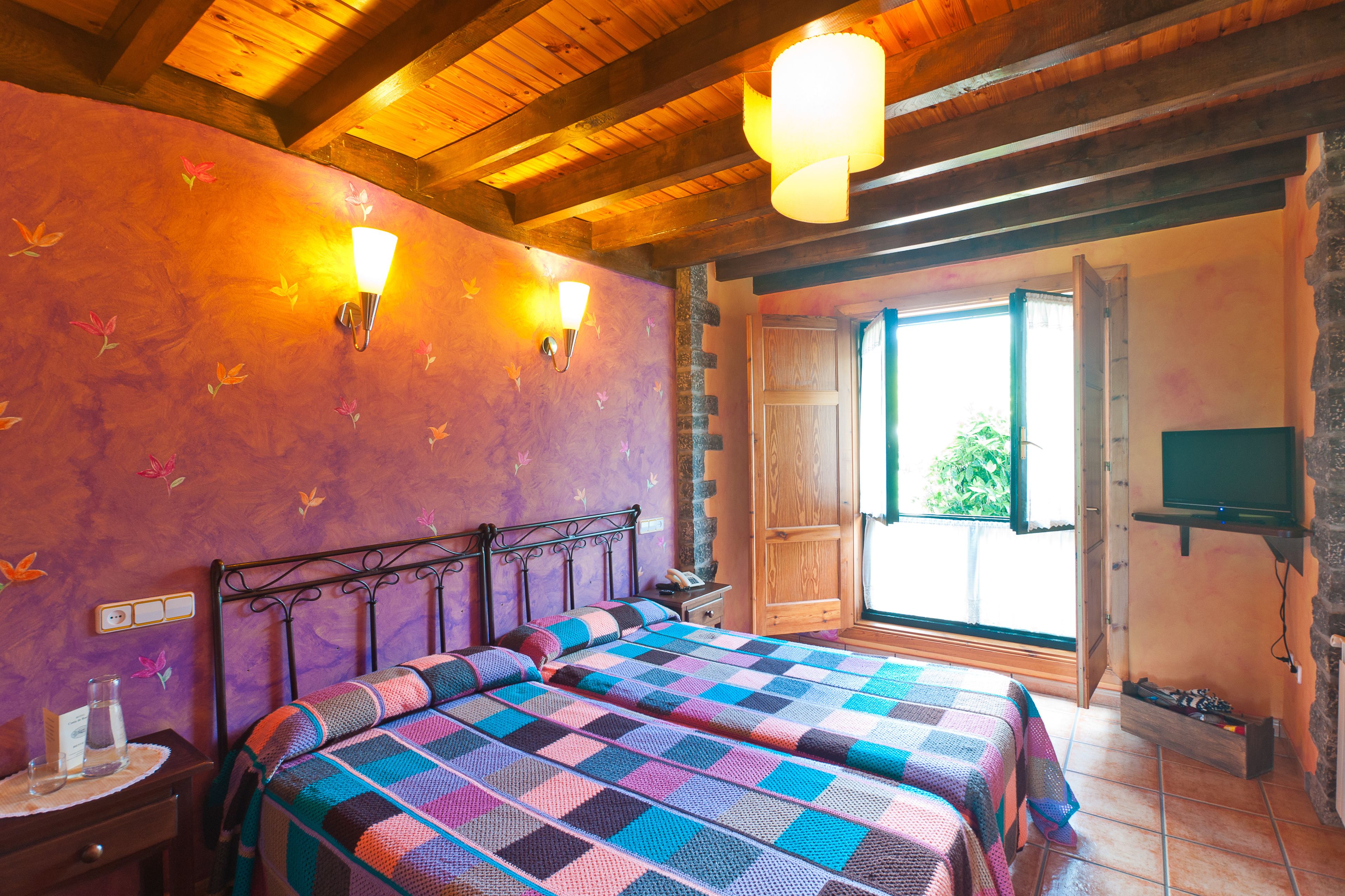 Une chambre avec un plafond en bois, deux lits recouverts de couvertures colorées, une table de chevet, un téléviseur fixé au mur et une fenêtre donnant sur un paysage verdoyant.