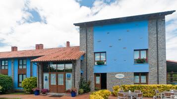 A hotel with a blue exterior, stone facade, and a tiled roof. There are outdoor seating areas with tables and chairs.
