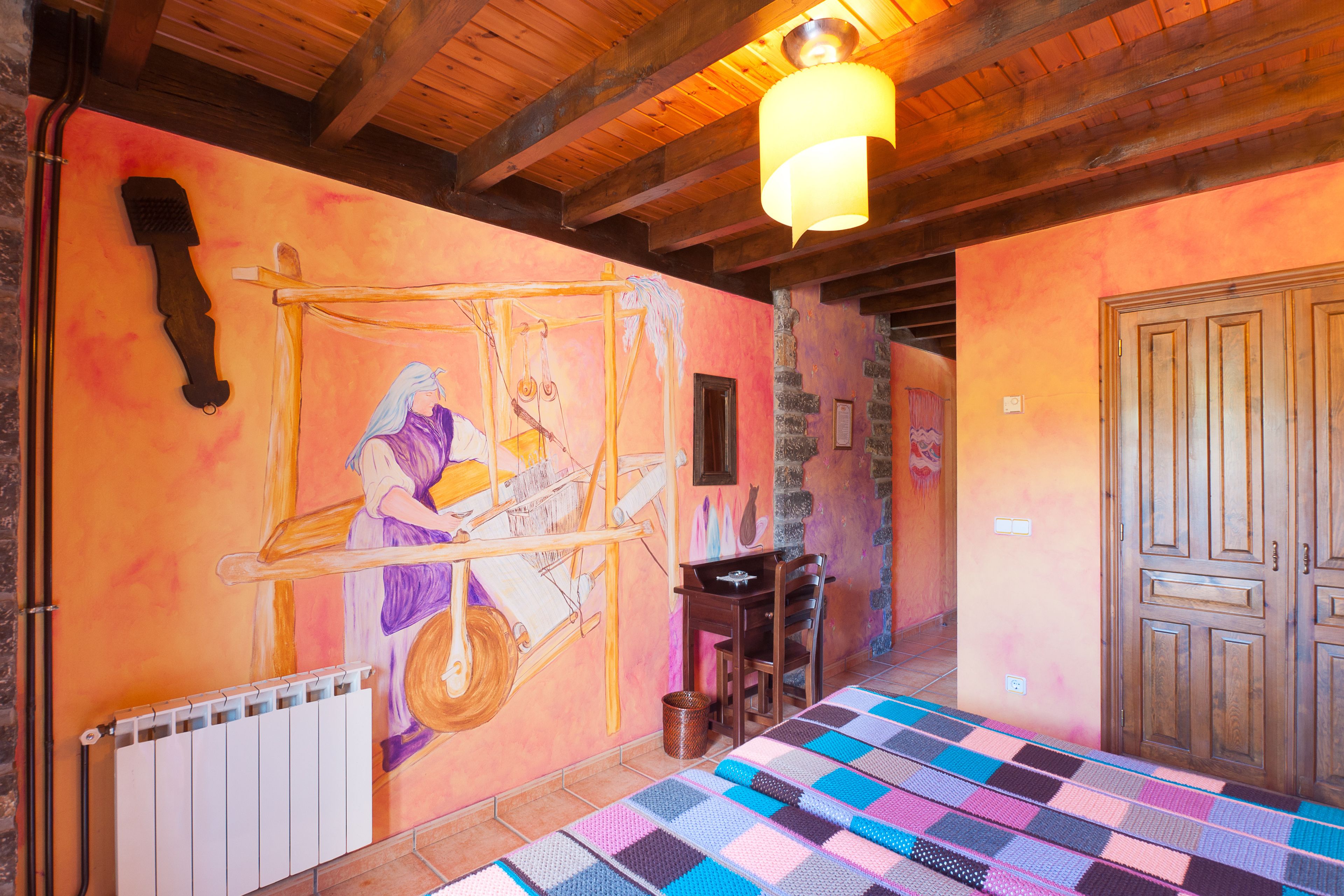A cozy room with a wooden ceiling, a colorful quilt on the floor, a mural on the wall, and a door to the right.