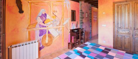 A cozy room with a wooden ceiling, a colorful quilt on the floor, a mural on the wall, and a door to the right.