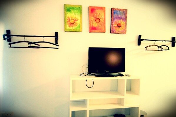 A room with a TV on a white stand, three paintings on the wall, and two black hairdryers on hooks.